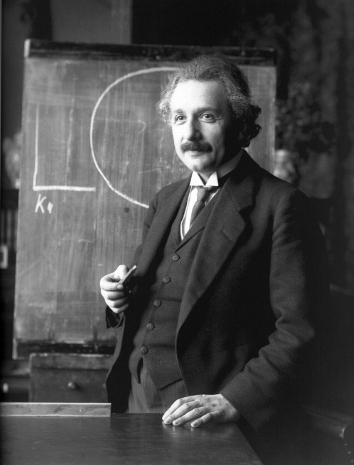 Albert Einstein en un traje negro elegante al lado de una pizarra.